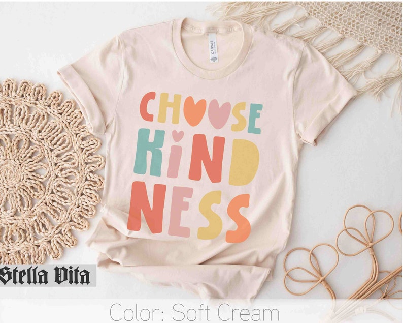 K&ouml;nnte beinhalten: Ein T-Shirt in zartem Creme mit dem Text "Choose Kindness" in bunten Blockbuchstaben mit Herzen.
