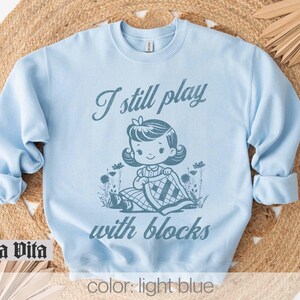 K&ouml;nnte beinhalten: Hellblauer Pullover mit dem Text "I still play with blocks" und einer Cartoon-Illustration eines M&auml;dchens, das mit Bl&ouml;cken spielt.