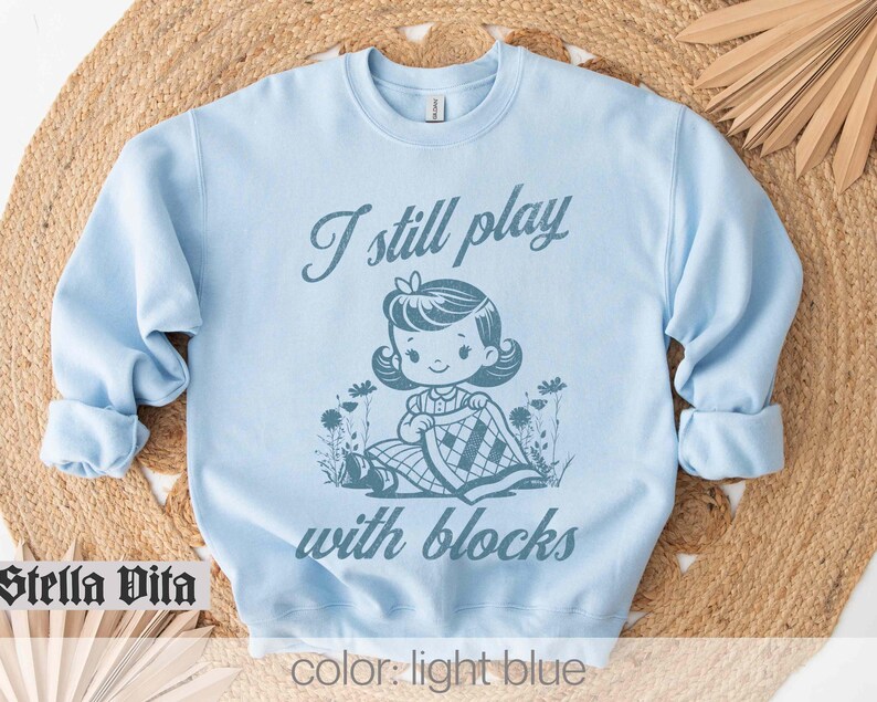 K&ouml;nnte beinhalten: Hellblauer Pullover mit dem Text "I still play with blocks" und einer Cartoon-Illustration eines M&auml;dchens, das mit Bl&ouml;cken spielt.