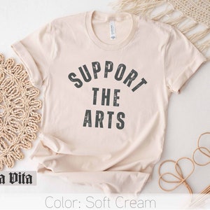 May include: A soft cream colored t-shirt with the text "SUPPORT THE ARTS" printed in black.