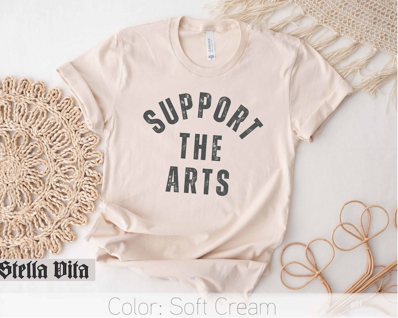 May include: A soft cream colored t-shirt with the text "SUPPORT THE ARTS" printed in black.