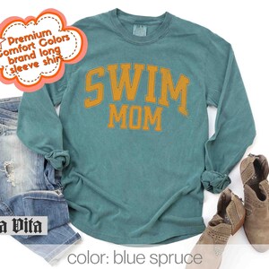 Puede incluir: Una camiseta de manga larga azul turquesa con el texto "SWIM MOM" en letras doradas. La camiseta est&aacute; hecha de una tela suave y c&oacute;moda.