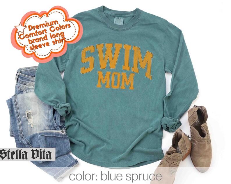 Puede incluir: Una camiseta de manga larga azul turquesa con el texto "SWIM MOM" en letras doradas. La camiseta est&aacute; hecha de una tela suave y c&oacute;moda.