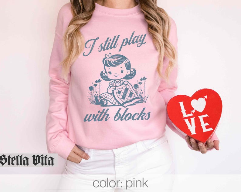 K&ouml;nnte beinhalten: Rosafarbener Pullover mit dem Text "I still play with blocks" und einem Cartoon-M&auml;dchen, das mit Bl&ouml;cken spielt. Der Pullover wird von einer Person getragen, die eine rote herzf&ouml;rmige Schachtel mit dem Wort "LOVE" h&auml;lt.