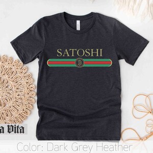 May include: Dark grey heather t-shirt with the word "SATOSHI" printed in white and a green, red, and gold stripe with a Bitcoin logo in the center.
