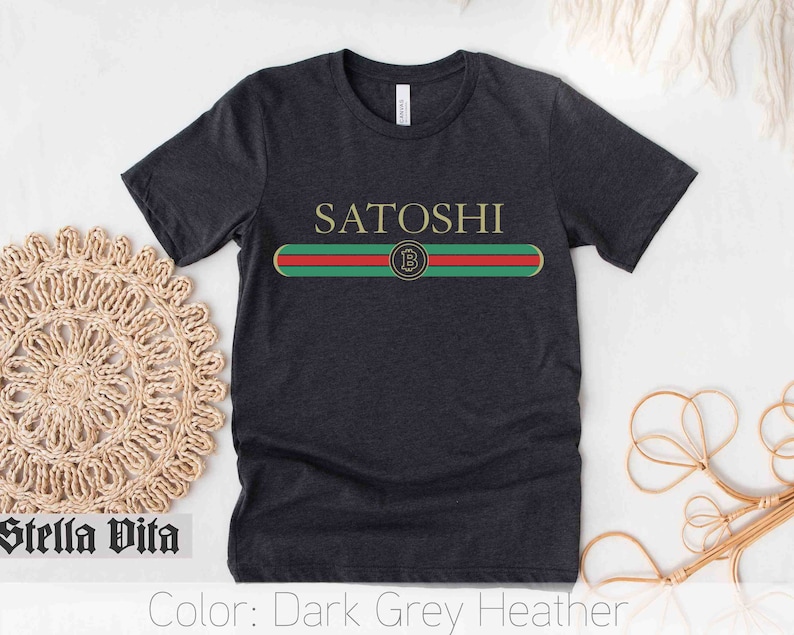 May include: Dark grey heather t-shirt with the word "SATOSHI" printed in white and a green, red, and gold stripe with a Bitcoin logo in the center.