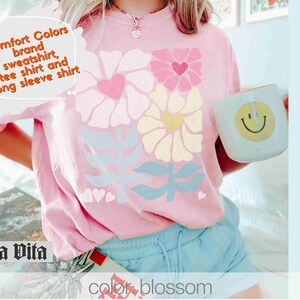 May include: A pink t-shirt with a floral design and the text "Comfort Colors brand sweatshirt, tee shirt and long sleeve shirt". The shirt is being worn by a person and has a blue and white floral design.