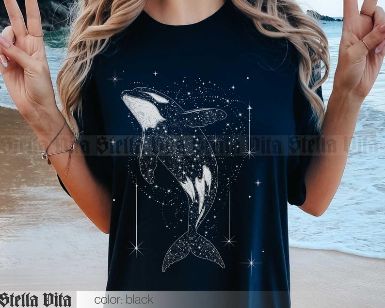 May include: A black t-shirt with a white graphic of an orca whale swimming through a field of stars. The orca is outlined in white and filled with a pattern of white stars.