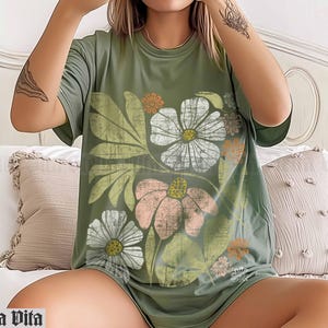 May include: A green t-shirt with a floral print featuring a variety of flowers in shades of pink, white, and yellow. The flowers are surrounded by green leaves and stems.