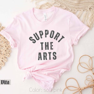 May include: A soft pink t-shirt with the text "SUPPORT THE ARTS" in black lettering.