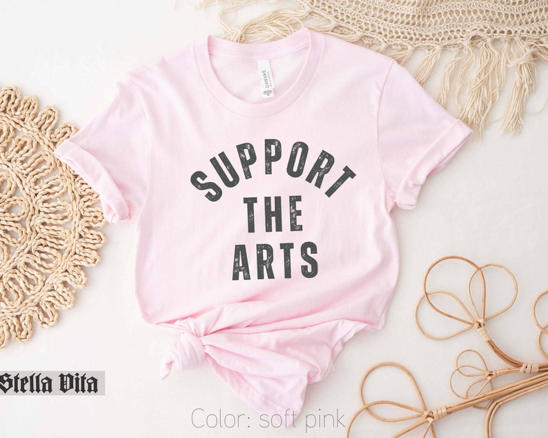 May include: A soft pink t-shirt with the text "SUPPORT THE ARTS" in black lettering.