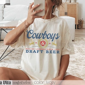 May include: Ivory-colored t-shirt with the text "Cowboys & Draft Beer" in blue and red. The shirt features a vintage-style design with the words "Making Legends Since 1988". The shirt is made of comfort colors material.