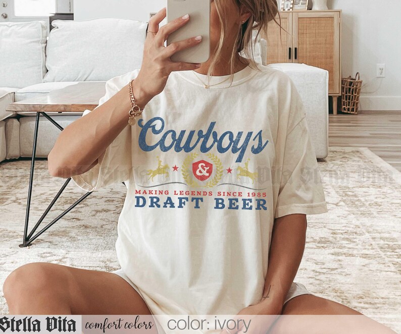 May include: Ivory-colored t-shirt with the text "Cowboys & Draft Beer" in blue and red. The shirt features a vintage-style design with the words "Making Legends Since 1988". The shirt is made of comfort colors material.