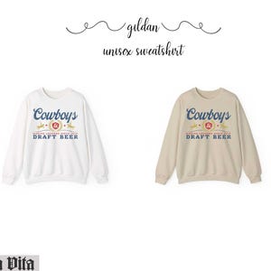 May include: Two Gildan unisex sweatshirts, one white and one beige. Each features the word "Cowboys" in blue script above a graphic and the words "DRAFT BEER" in blue. The text "MAKING LEGENDS SINCE 1912" is also visible.