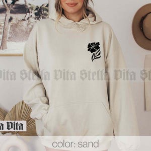 May include: A sand-colored hoodie with a black floral design on the left chest. The hoodie has a drawstring hood and a kangaroo pocket.