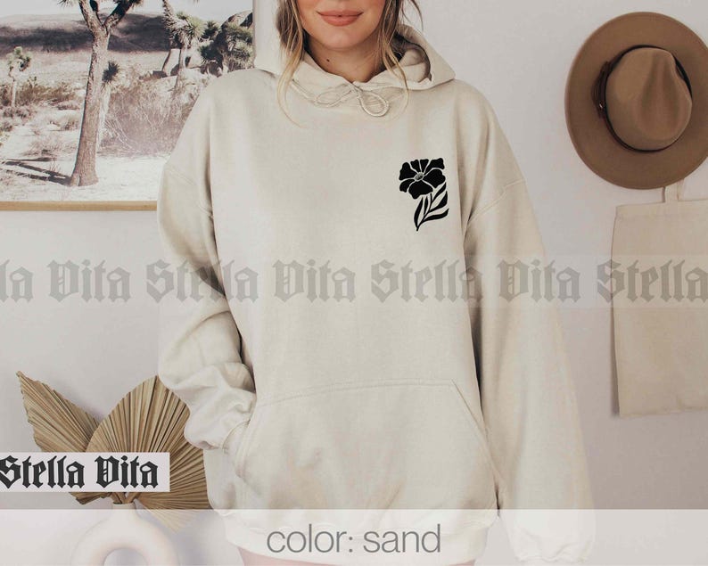 May include: A sand-colored hoodie with a black floral design on the left chest. The hoodie has a drawstring hood and a kangaroo pocket.