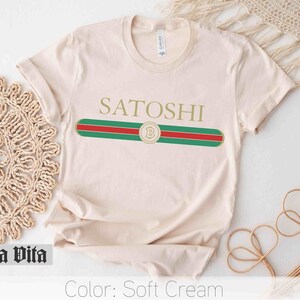 May include: A soft cream colored t-shirt with a gold "Satoshi" text and a green, red, and gold stripe design with a gold "B" inside a circle.