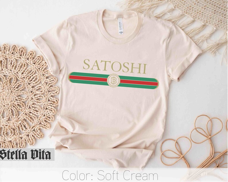 May include: A soft cream colored t-shirt with a gold "Satoshi" text and a green, red, and gold stripe design with a gold "B" inside a circle.