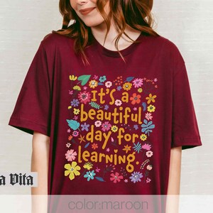 May include: A maroon t-shirt with a floral design and the text "It's a beautiful day for learning."