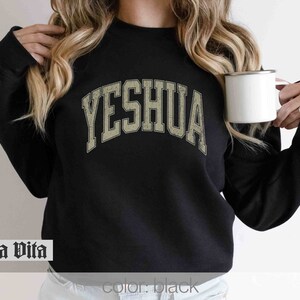 May include: Black sweatshirt with a vintage-style, arched graphic that reads "YESHUA" in a beige color.