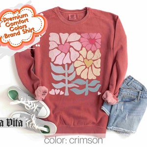 May include: Crimson red sweatshirt with a floral design featuring hearts and leaves. The design is in shades of pink, yellow, blue, and green.