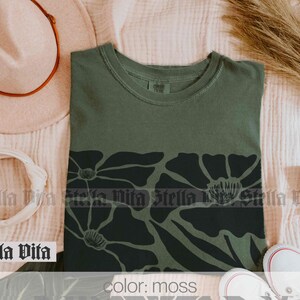 Puede incluir: Una camiseta verde musgo con un estampado floral negro. El estampado presenta flores grandes y estilizadas con un c&iacute;rculo central.