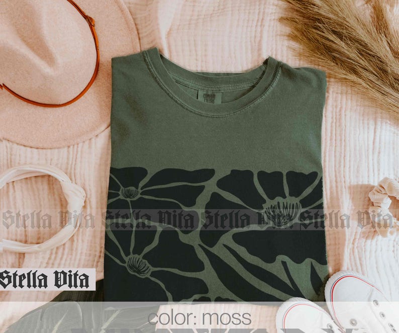 Puede incluir: Una camiseta verde musgo con un estampado floral negro. El estampado presenta flores grandes y estilizadas con un c&iacute;rculo central.
