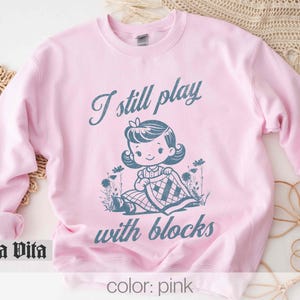 K&ouml;nnte beinhalten: Rosafarbener Pullover mit dem Text "I still play with blocks" und einer Cartoon-Illustration eines M&auml;dchens, das mit Bl&ouml;cken spielt.