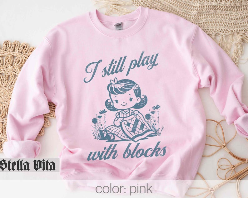 K&ouml;nnte beinhalten: Rosafarbener Pullover mit dem Text "I still play with blocks" und einer Cartoon-Illustration eines M&auml;dchens, das mit Bl&ouml;cken spielt.