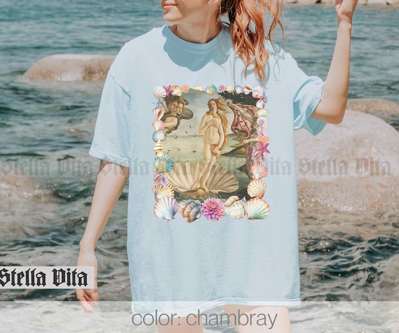 Puede incluir: Una camiseta azul claro con un estampado de una pintura de una mujer de pie sobre una concha de mar, rodeada de conchas y flores. El texto "color: chambray" est&aacute; en la parte inferior de la imagen.