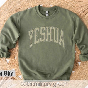May include: A military green sweatshirt with the word "YESHUA" printed in a large, white, arched font.