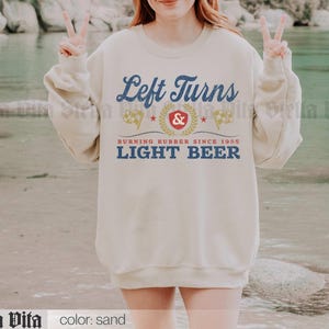 May include: Sand-colored sweatshirt with the text "Left Turns Light Beer" in blue and white. The design includes a red emblem with a star and laurel wreath, and the phrase "Burning Rubber Since 1985."