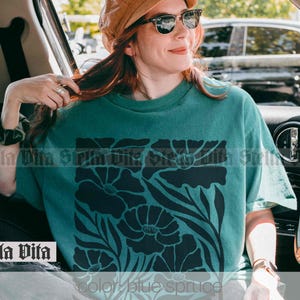 Puede incluir: Una camiseta azul turquesa con un dise&ntilde;o floral negro. El dise&ntilde;o presenta flores grandes con detalles intrincados y tallos. La camiseta tiene un corte holgado y cuello redondo.