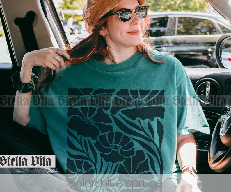Puede incluir: Una camiseta azul turquesa con un dise&ntilde;o floral negro. El dise&ntilde;o presenta flores grandes con detalles intrincados y tallos. La camiseta tiene un corte holgado y cuello redondo.