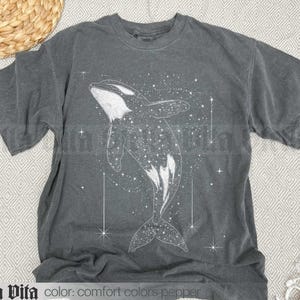 May include: A dark grey t-shirt featuring a white whale silhouette design. The whale is surrounded by white stars and a galaxy-like pattern.