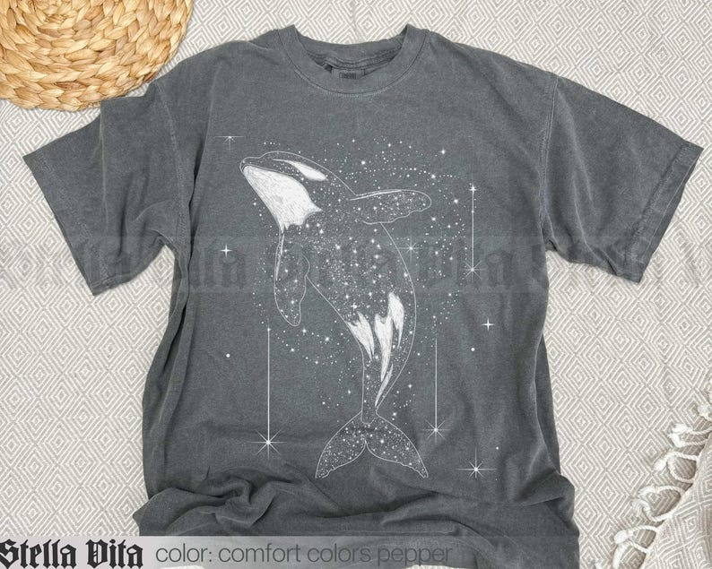 May include: A dark grey t-shirt featuring a white whale silhouette design. The whale is surrounded by white stars and a galaxy-like pattern.
