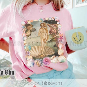 Puede incluir: Una camiseta rosa con un estampado gr&aacute;fico de la pintura El nacimiento de Venus rodeado de conchas marinas. El texto "color: blossom" est&aacute; impreso en la parte inferior de la imagen.