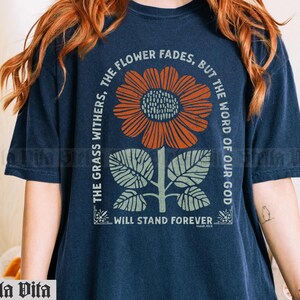 May include: A navy blue t-shirt with a graphic of a sunflower and leaves. The text "The grass withers, the flower fades, but the word of our God will stand forever" is printed on the shirt.