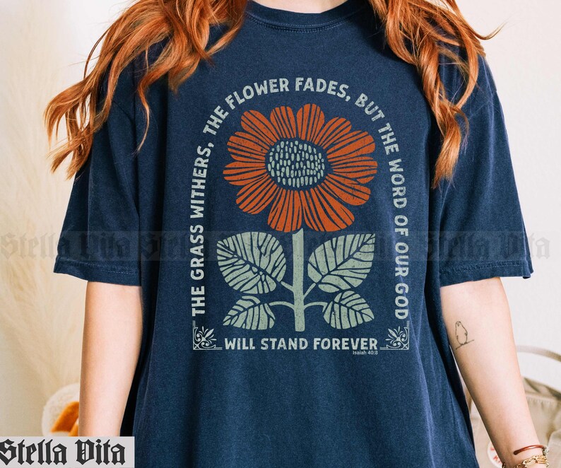 May include: A navy blue t-shirt with a graphic of a sunflower and leaves. The text "The grass withers, the flower fades, but the word of our God will stand forever" is printed on the shirt.