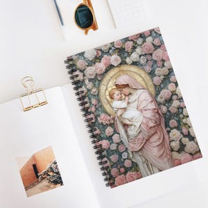 Puede incluir: Una libreta de espiral con un diseño floral rosa y blanco. La portada presenta una pintura de una mujer sosteniendo un bebé. La mujer lleva una túnica rosa y el bebé lleva una túnica blanca. La pintura está rodeada por un halo de luz.