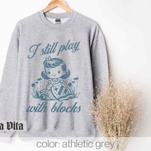 K&ouml;nnte beinhalten: Ein graues Sweatshirt mit dem Text "I still play with blocks" in Blau. Der Text wird von einer Retro-Illustration eines M&auml;dchens begleitet, das mit Bl&ouml;cken spielt.