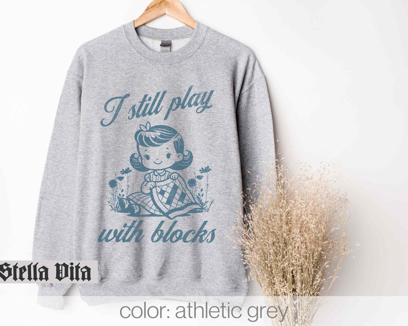 K&ouml;nnte beinhalten: Ein graues Sweatshirt mit dem Text "I still play with blocks" in Blau. Der Text wird von einer Retro-Illustration eines M&auml;dchens begleitet, das mit Bl&ouml;cken spielt.