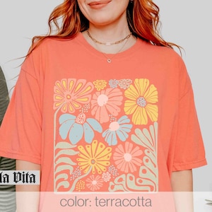 Peut inclure: Un t-shirt de couleur terre cuite avec un imprim&eacute; graphique de fleurs color&eacute;es dans une forme carr&eacute;e. Le texte "Stella Vita" est visible dans le coin inf&eacute;rieur gauche de l'image.