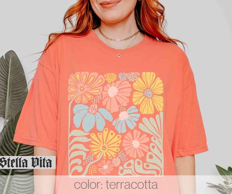 Peut inclure: Un t-shirt de couleur terre cuite avec un imprim&eacute; graphique de fleurs color&eacute;es dans une forme carr&eacute;e. Le texte "Stella Vita" est visible dans le coin inf&eacute;rieur gauche de l'image.