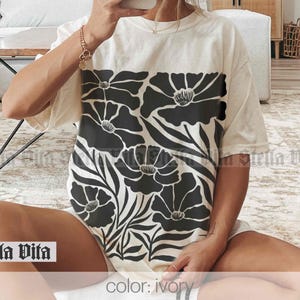 Puede incluir: Una camiseta blanca con un estampado floral negro. El estampado presenta flores grandes y estilizadas con hojas. La camiseta est&aacute; hecha de una tela suave y c&oacute;moda y tiene un corte holgado. El texto "Stella Vita" es visible en la parte inferior de la imagen. El color de la camiseta es marfil.