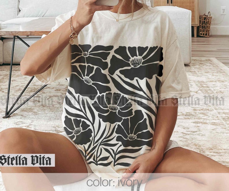 Puede incluir: Una camiseta blanca con un estampado floral negro. El estampado presenta flores grandes y estilizadas con hojas. La camiseta est&aacute; hecha de una tela suave y c&oacute;moda y tiene un corte holgado. El texto "Stella Vita" es visible en la parte inferior de la imagen. El color de la camiseta es marfil.