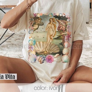 Puede incluir: Una camiseta blanca con un estampado gr&aacute;fico de una mujer de pie sobre una concha. El gr&aacute;fico est&aacute; rodeado de conchas y flores. El texto "Stella Vita" est&aacute; impreso en la esquina inferior izquierda de la camiseta. El texto "color: ivory" est&aacute; impreso en la esquina inferior derecha de la camiseta.