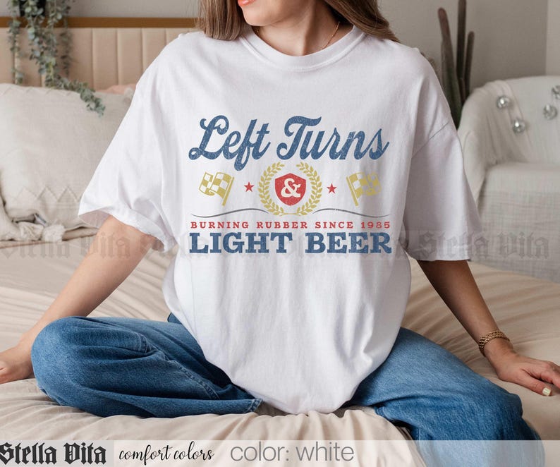 May include: White t-shirt with the text "Left Turns" in blue script above a graphic with flags and the words "Burning Rubber Since 1985 Light Beer". The shirt is a comfort color.