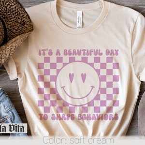 May include: A soft cream colored t-shirt with a pink checkered pattern and a pink smiley face with hearts for eyes. The text "It's a beautiful day to shape behaviors" is printed on the shirt.