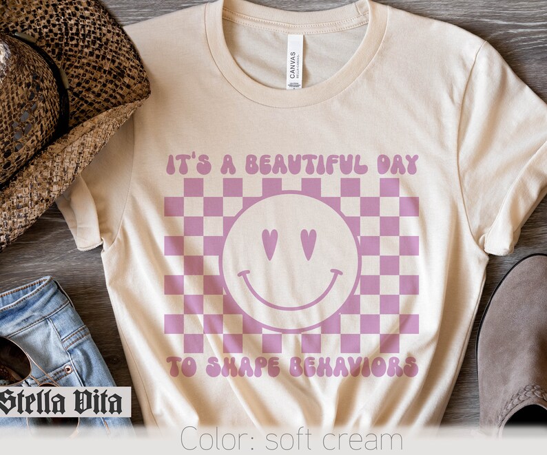 May include: A soft cream colored t-shirt with a pink checkered pattern and a pink smiley face with hearts for eyes. The text "It's a beautiful day to shape behaviors" is printed on the shirt.